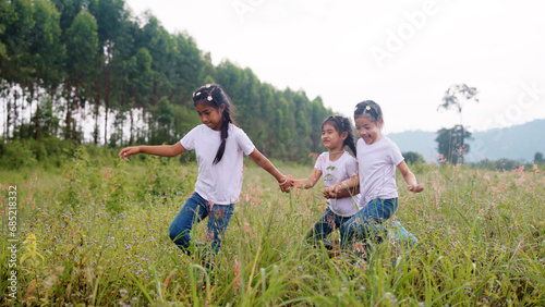 Three active sister cute child girls hold hands move way forward go walk in rural green grass field. Happy sibling young kids group asia people relax smile joy explore fun in love care nature forest.