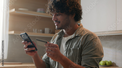 Happy Arabian man with phone and credit card using instant mobile payment at home kitchen Indian guy male customer shopper buyer buying online purchase smartphone shopping food delivery e-banking app photo