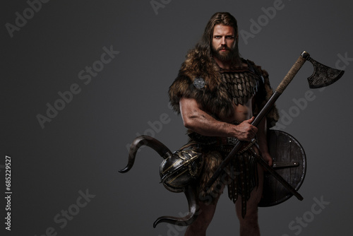 A fearless Viking warrior with a majestic beard adorned in fur and light armor, carrying an axe and shield, standing against a gray background