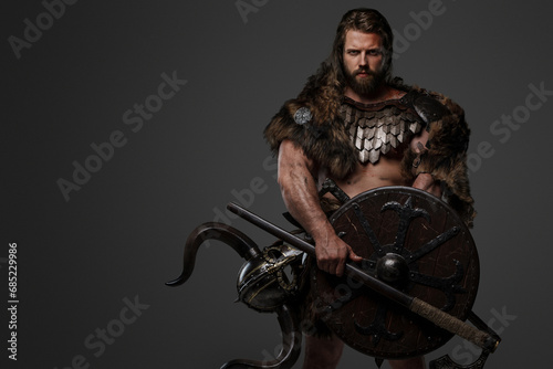 A fearless Viking warrior with a majestic beard adorned in fur and light armor, carrying an axe and shield, standing against a gray background