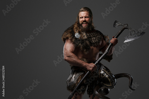 Fierce, bearded Viking warrior dressed in fur and light armor, holding a large two-handed axe, shouting against a grey background