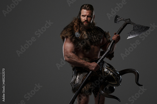 A fierce bearded Viking warrior in fur and light armor, with a helmet attached to his belt, holding a large two-handed axe on a gray background