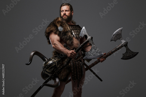 A fierce bearded Viking warrior in fur and light armor, with a helmet attached to his belt, holding a large two-handed axe on a gray background