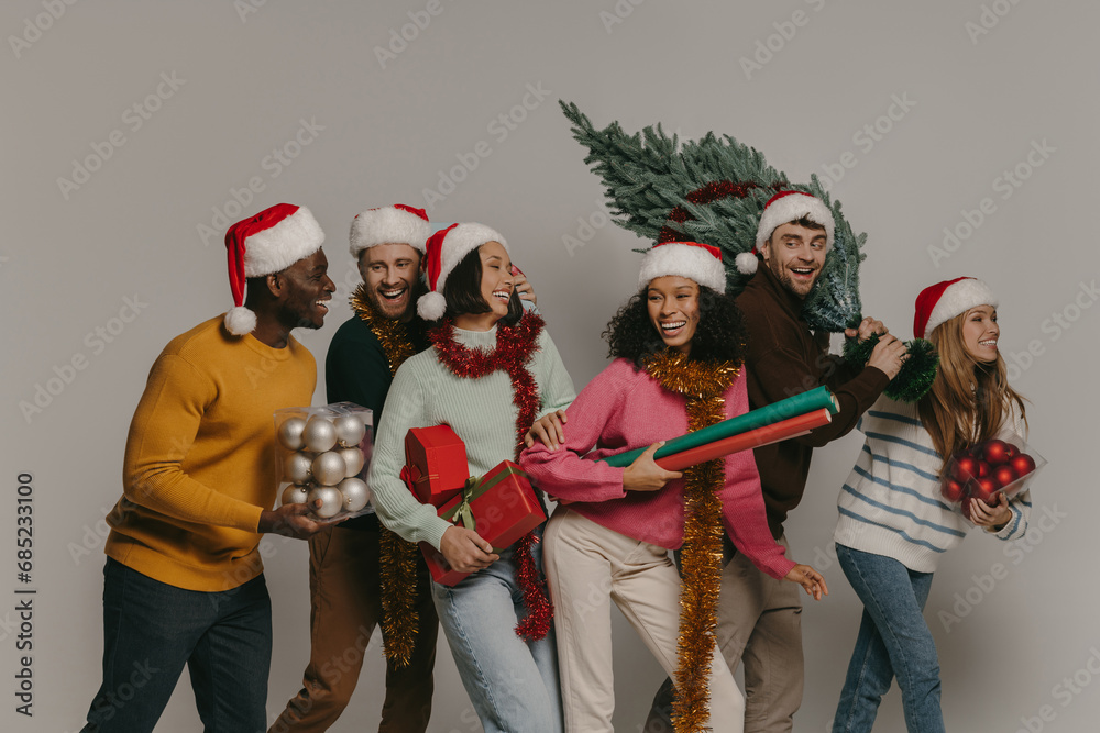 Happy young people carrying Christmas tree and various ornaments while walking on studio background Happy young people carrying Christmas tree and various ornaments while walking on studio background