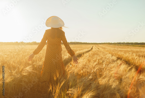 Beautiful young woman walks across the field and touches rye with her hand. Girl in the rays of the sunset. Freedom and happiness concept.
