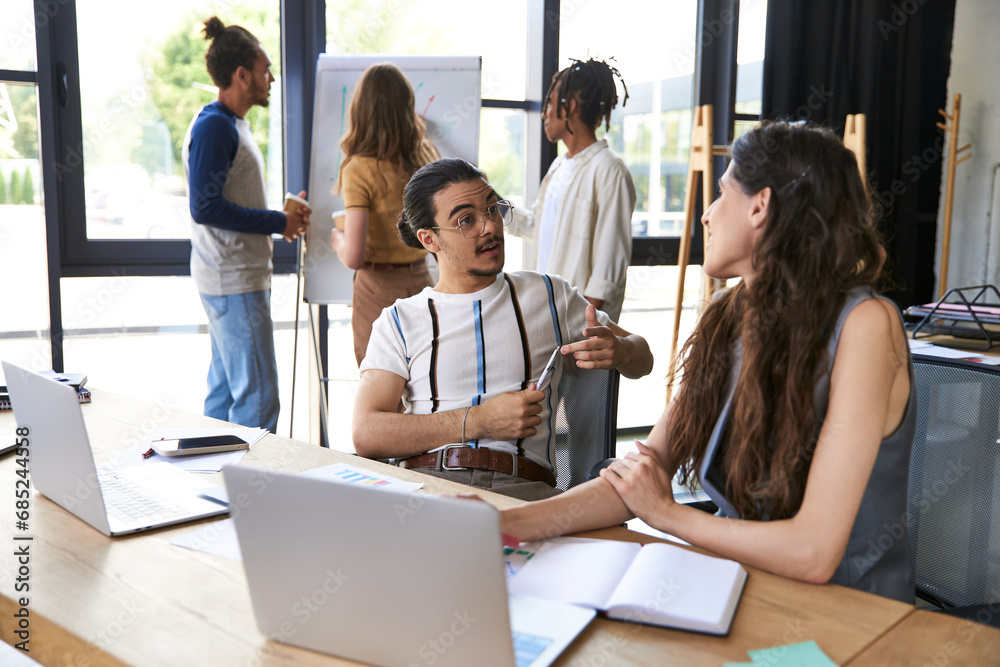 young businessman in eyeglasses talking to female colleague near multicultural team in office young businessman in eyeglasses talking to female colleague near multicultural team in office