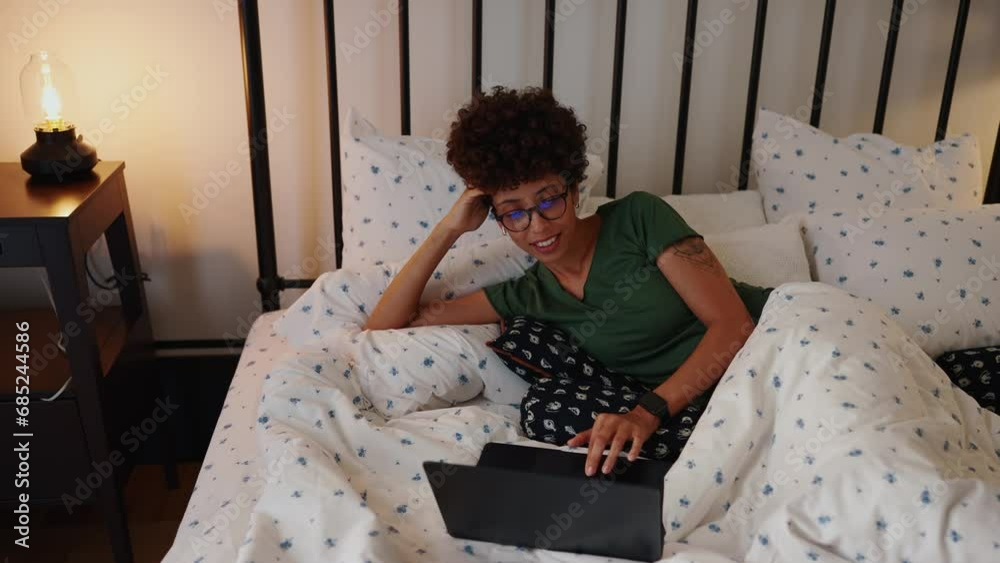 Beautiful african woman with glasses and curly hair working on laptop on bed from home Beautiful african woman with glasses and curly hair working on laptop on bed from home
