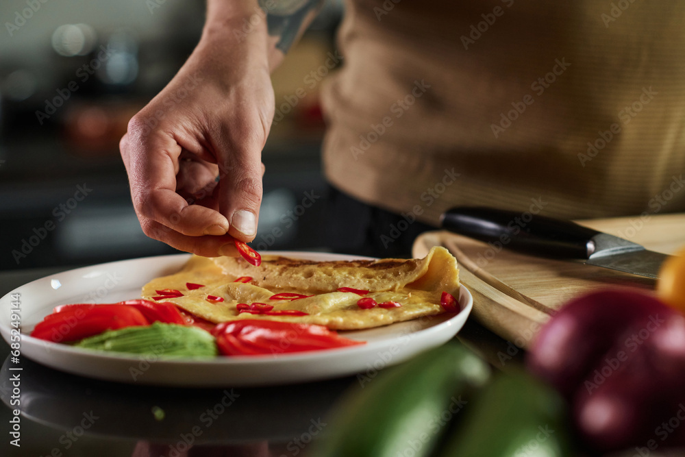 Closeup of hand of unrecognizable man placing slice of chili pepper on omelet