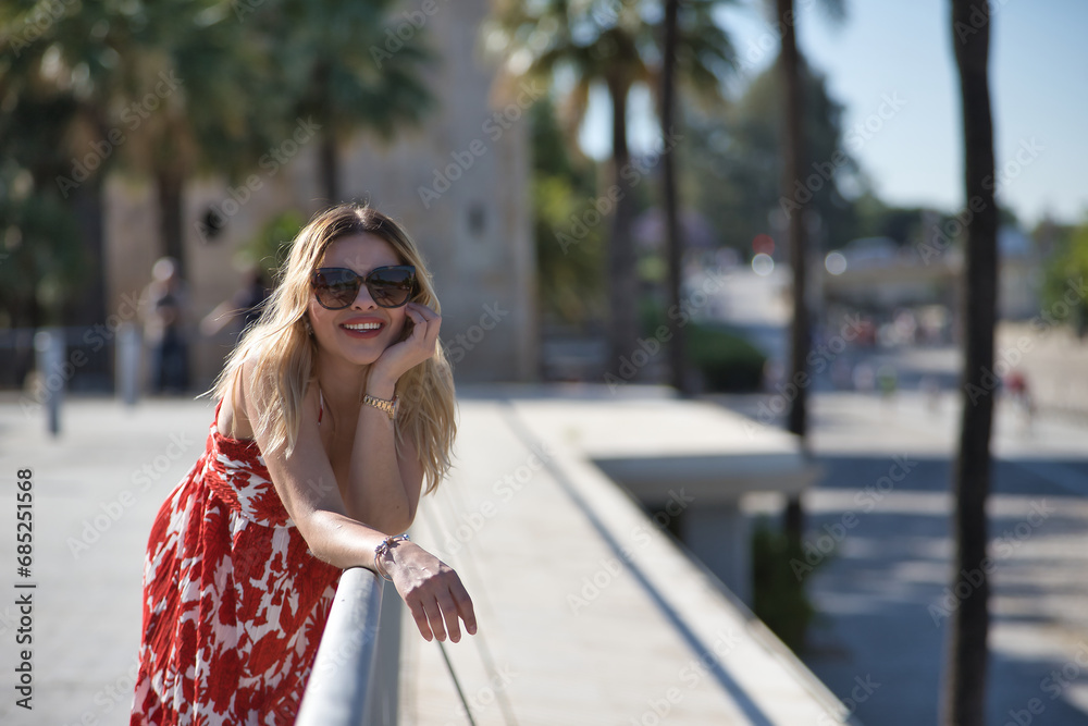 Obraz premium South American woman, young, beautiful, blonde with flower dress and sunglasses, leaning on a railing looking at camera smiling and happy. Concept travel, destinations, vacation.