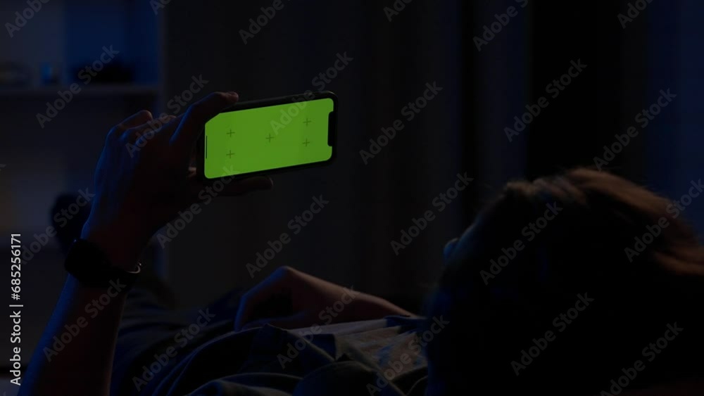 A man lying on the couch at night and holding horizontally a smartphone with a green screen. The man is watching a video, movie or series. Advertising area, workplace. A man lying on the couch at night and holding horizontally a smartphone with a green screen. The man is watching a video, movie or series. Advertising area, workplace.