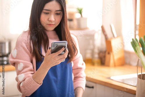 Pretty woman looking at message on mobile phone at her kitchen, reads information from a smartphone