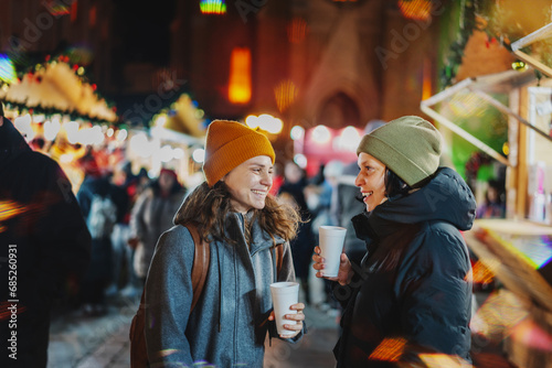Two happy young women friends with mulled wine stands walking at a Christmas market in a European city photo