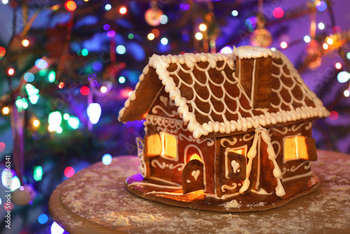 Big gingerbread house on wooden table, flour snow around, defocused colorful lights of Christmas decorated fir tree background. Night holiday illumination. Merry Xmas and Happy New Year Greeting card.