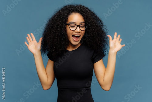 Foto Joyful young woman with glasses and curly hair, excitedly raising hands, on blue background