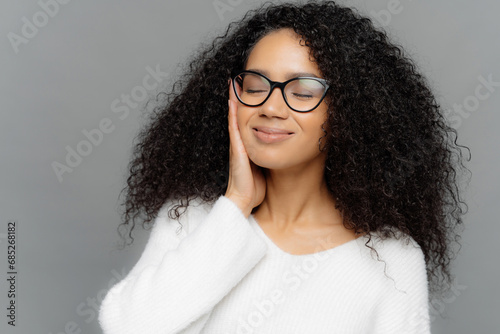 Serene woman with glasses and curly hair, hand on cheek, eyes closed, on a grey background, exuding calm