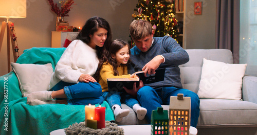 Happy Caucasian family with small kid sitting at cozy decorated room in festive mood reading book together on Christmas Eve. Happy holidays. New Year celebration concept. Winter holidays season