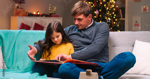 Portrait of handsome Caucasian father reading a book of fairytales for his pretty little daughter at decorated home on Christmas night. New Year celebration. Family concept. Magic moments