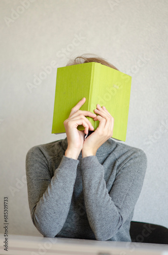 A portrait of a woman in a gray sweater covers her face with a green diary. The agony of time management and planning. The concept of challenges and problems.