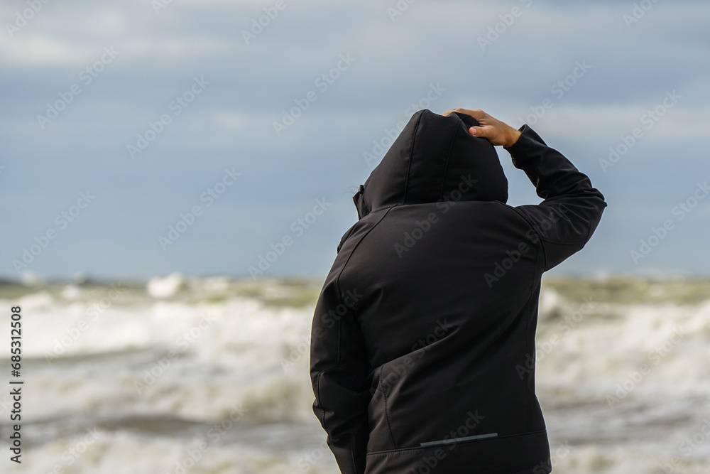 A man in a black jacket with a hood holds the hood and looks at the stormy sea, rear view A man in a black jacket with a hood holds the hood and looks at the stormy sea, rear view