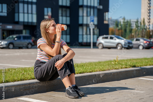  A young girl is sitting on the sidewalk and drinking coffee, a city street. Togliatti, Russia - 3 Sep 2023