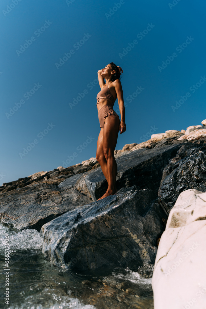 A beautiful young brunette woman in a swimsuit stands on the rocks by the sea.