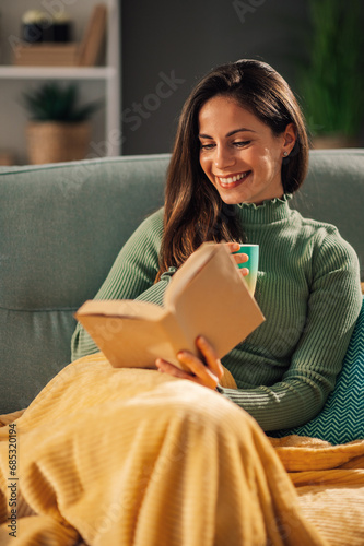 Smiling woman relaxing on a couch at home covered in blankets reading.