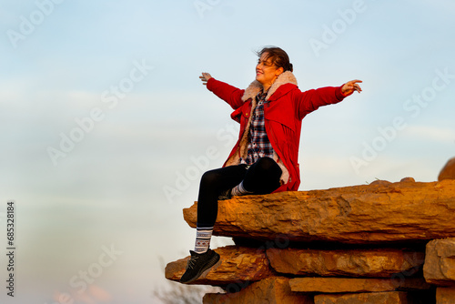 Beautiful Caucasian woman sit on the rock near cliff on the mountain and spread her arms with happiness and look forward with warm light in the morning.