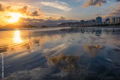 City of Santos, Brazil. Sunset on Santos beach, sky reflected in the wet sand. Seaside buildings lit by the sun against the blue sky.