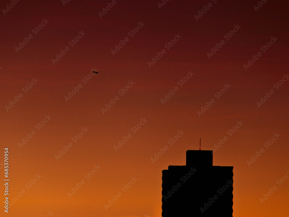 Minimalist image with the silhouette of an apartment building and a jet plane crossing the horizon during twilight.