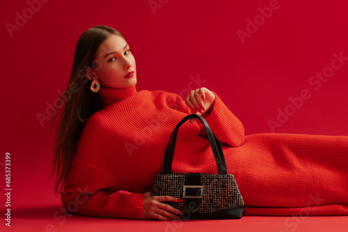 Fashionable confident woman wearing trendy red knitted turtleneck dress, golden earrings, holding baguette bag, posing, laying on red background. Studio fashion portrait. Copy, empty space for text