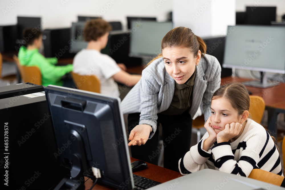Caring young female teacher calming upset teenage schoolgirl sitting at computer in classroom..