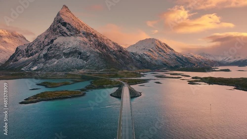 Aerial view of Fredvang bridge, sea and snowy mountains at colorful sunset in Lofoten Islands, Norway. Landscape with beautiful road, azure water, rocks in snow, sky with pink clouds. Top drone view