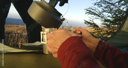 Woman sits in tent and keeps two cups with tea in hands. Tourist stands with boiling kettle and pours water in cups. Backpacker family warm up at cold windy weather on mountain valley. Hands close up. photo