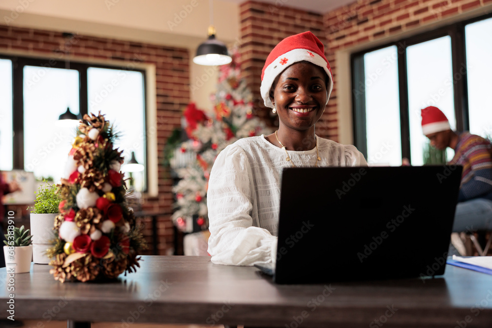 Smiling carefree african american employee in santa hat working on laptop in festive corporate environment portrait. Cheerful worker using computer at christmas decorated office