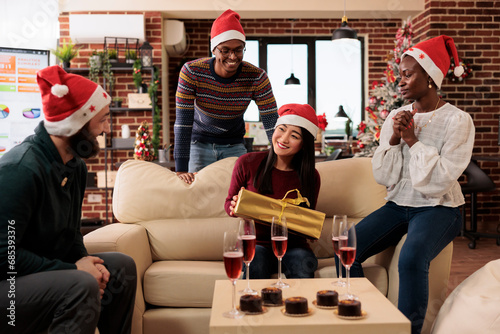 Asian woman employee in santa hat sitting on office couch and holding xmas gift box from colleagues. Diverse coworkers playing secret santa and sharing presents at new year holiday party