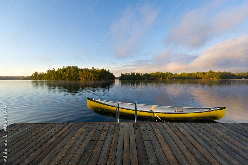 A yellow canoe is securely fastened to a rustic wooden dock nestled in the serene beauty of Muskoka, Ontario, Canada. In the distance, quaint cottages dot the landscape across the tranquil lake.