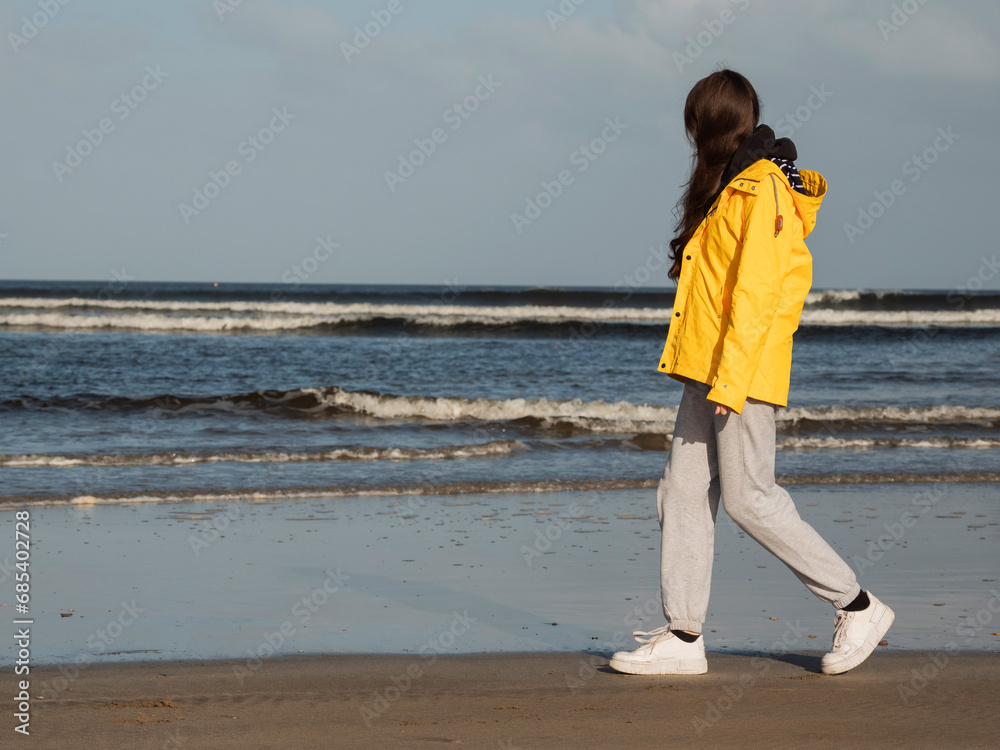 Teenager girl walking on a beach looking at the ocean. Young model in yellow jacket enjoying outdoor activity on fresh air. Selective focus. Cloudy sky. Travel and tourism concept.