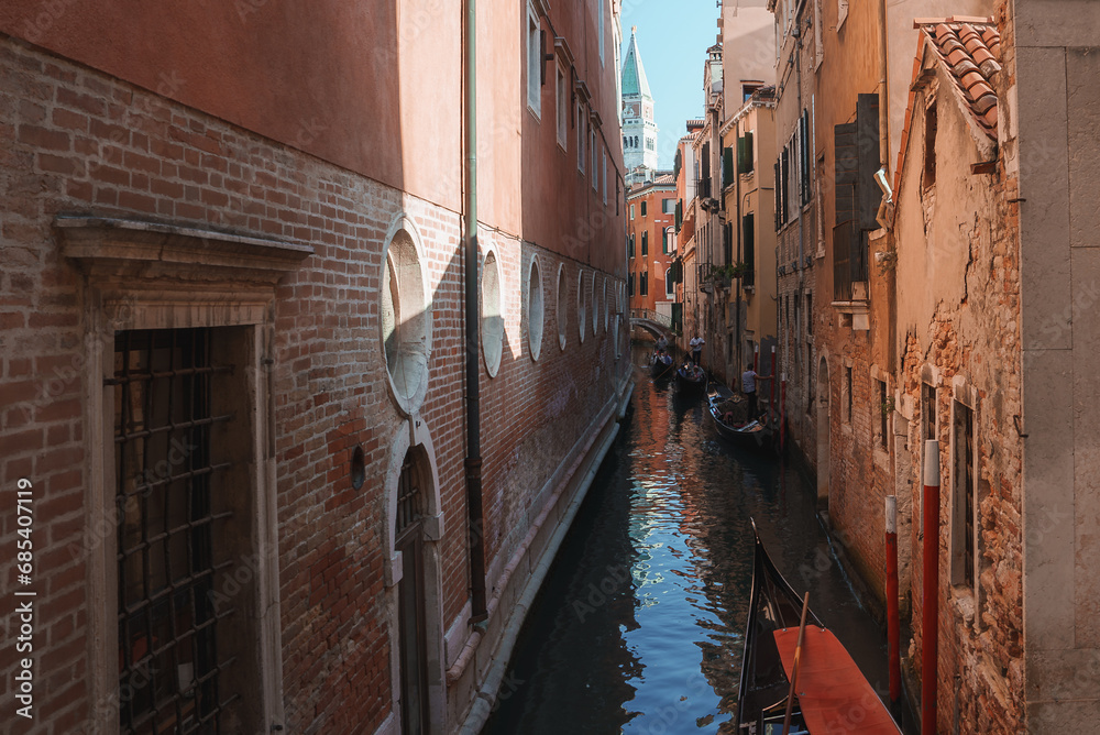 Scenic view of a narrow canal in Venice, Italy, surrounded by traditional reddish-brown buildings. Clear and sunny weather creates a peaceful and timeless atmosphere.
