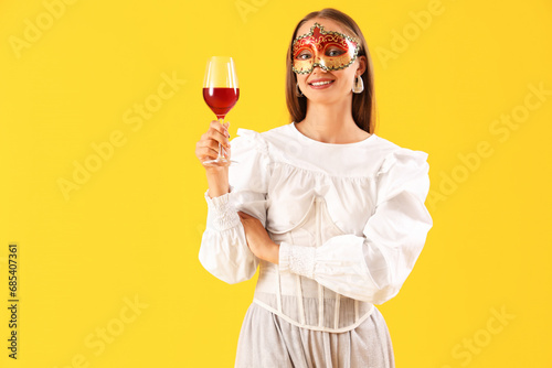 Beautiful young woman in carnival mask with glass of wine on yellow background photo