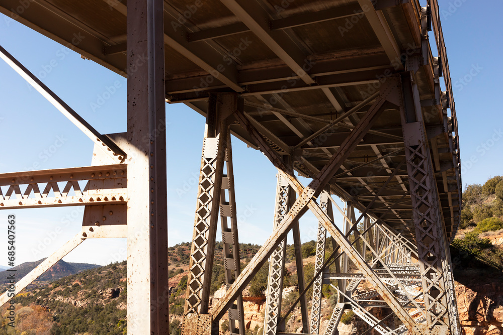 Fototapeta premium View Beneath Metal Construction Of Bridge Supports Against Blue Sky And Rocks. Rivets And Braces On Metal Beams. Midgley Bridge, Sedona Arizona. Horizontal Plane. Oak Creek Canyon, Coconino County