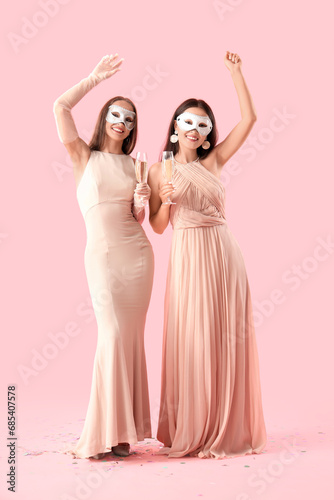 Beautiful young women in carnival masks with glasses of champagne on pink background
