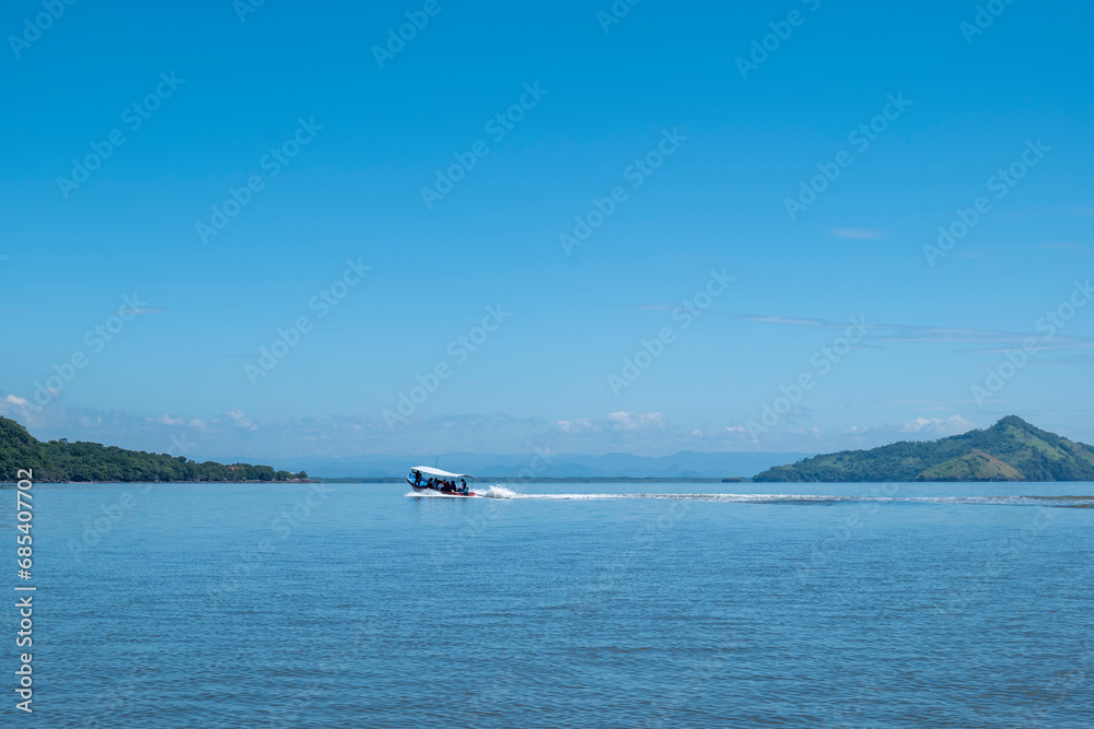 Speed Boat Rides on the Ocean between Coastlines Seen From Afar with a Volcanic Mountain, Lush Vegetation Forest, Beach, Houses and a Speed Boat on a Calm Ocean