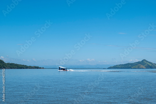 Obraz na płótnie Speed Boat Rides on the Ocean between Coastlines Seen From Afar with a Volcanic Mountain, Lush Vegetation Forest, Beach, Houses and a Speed Boat on a Calm Ocean w salonie