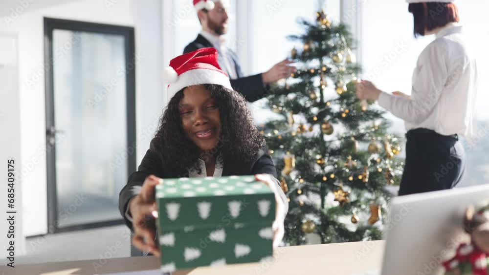 Successful adult businesswoman in Santa hat sitting at table holding gift box while colleagues decorating Christmas tree in modern office. Successful adult businesswoman in Santa hat sitting at table holding gift box while colleagues decorating Christmas tree in modern office.