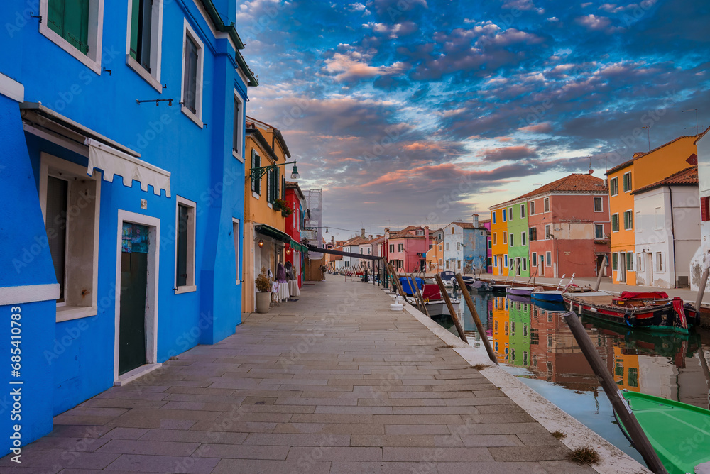Scenic view of colorful buildings along a waterway in Burano, Italy. Reflections of the buildings in the water. No people or activity visible. Unique architectural details not specified.