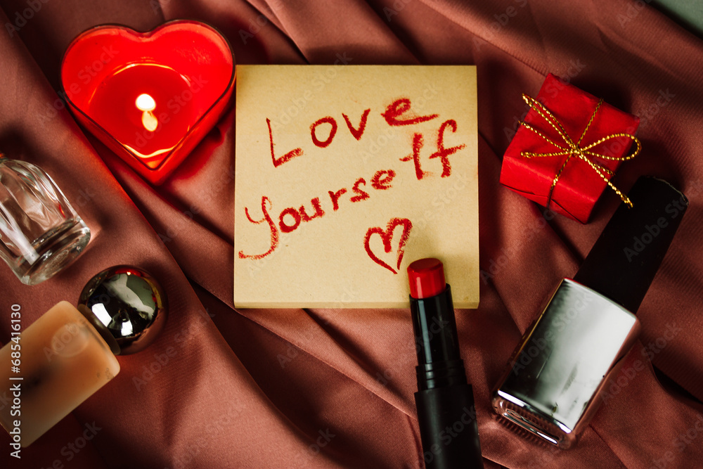 A note with a call to LOVE YOURSELF first drawing with red lipstick on paper for notes on silky pink cloth. A burning heart-shaped red candle, gift box, cosmetic bottles. Happy Valentines Day 2024. - obrazy, fototapety, plakaty 