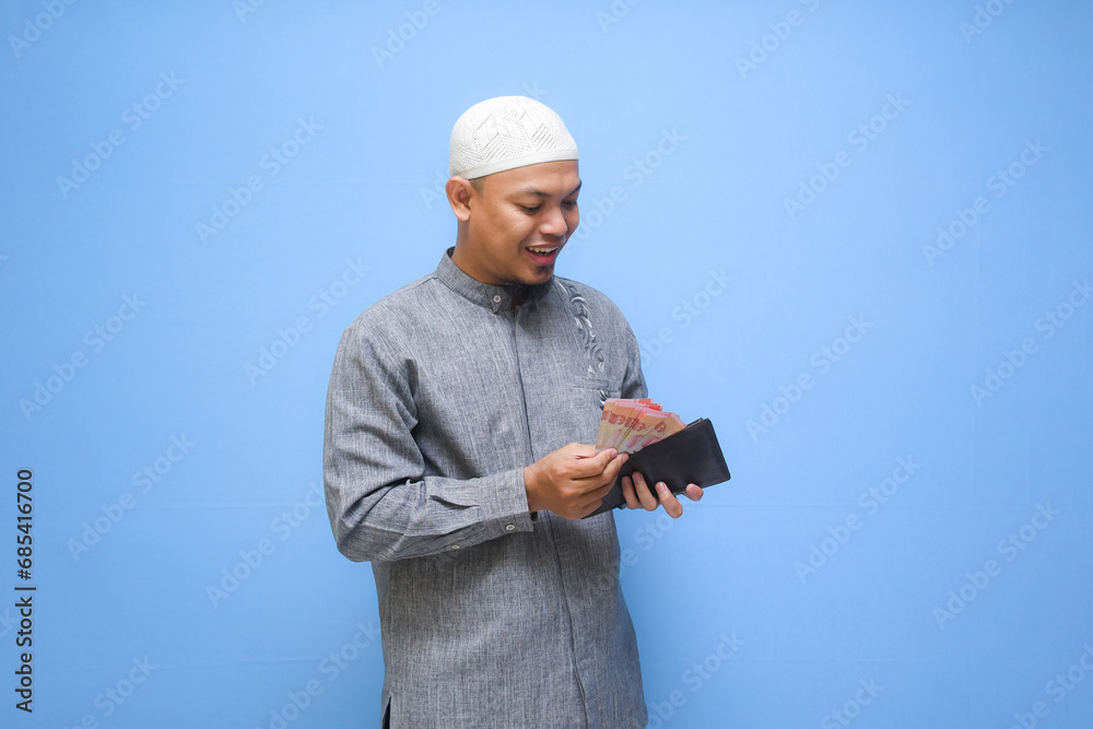 Asian Muslim man holding wallet of full cash money ready to pay zakat isolated on blue background. Islamic lifestyle concept Asian Muslim man holding wallet of full cash money ready to pay zakat isolated on blue background. Islamic lifestyle concept