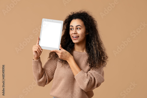 Young African-American woman holding tablet computer with blank screen on beige background