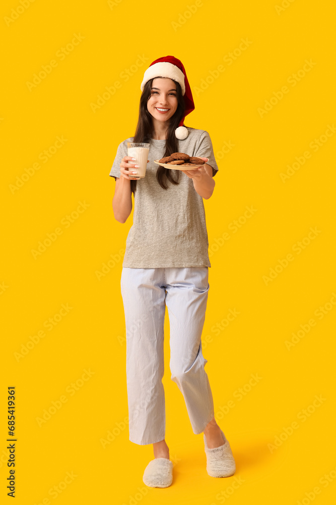 Beautiful young woman in pajamas with sweet cookies and glass of milk on yellow background