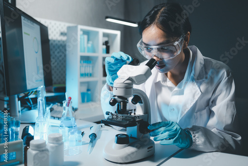 Young scientists in sterile clothing and safety goggles sitting at table conducting research investigations in a medical laboratory using a microscope, Serious concentrated female microbiologist. photo
